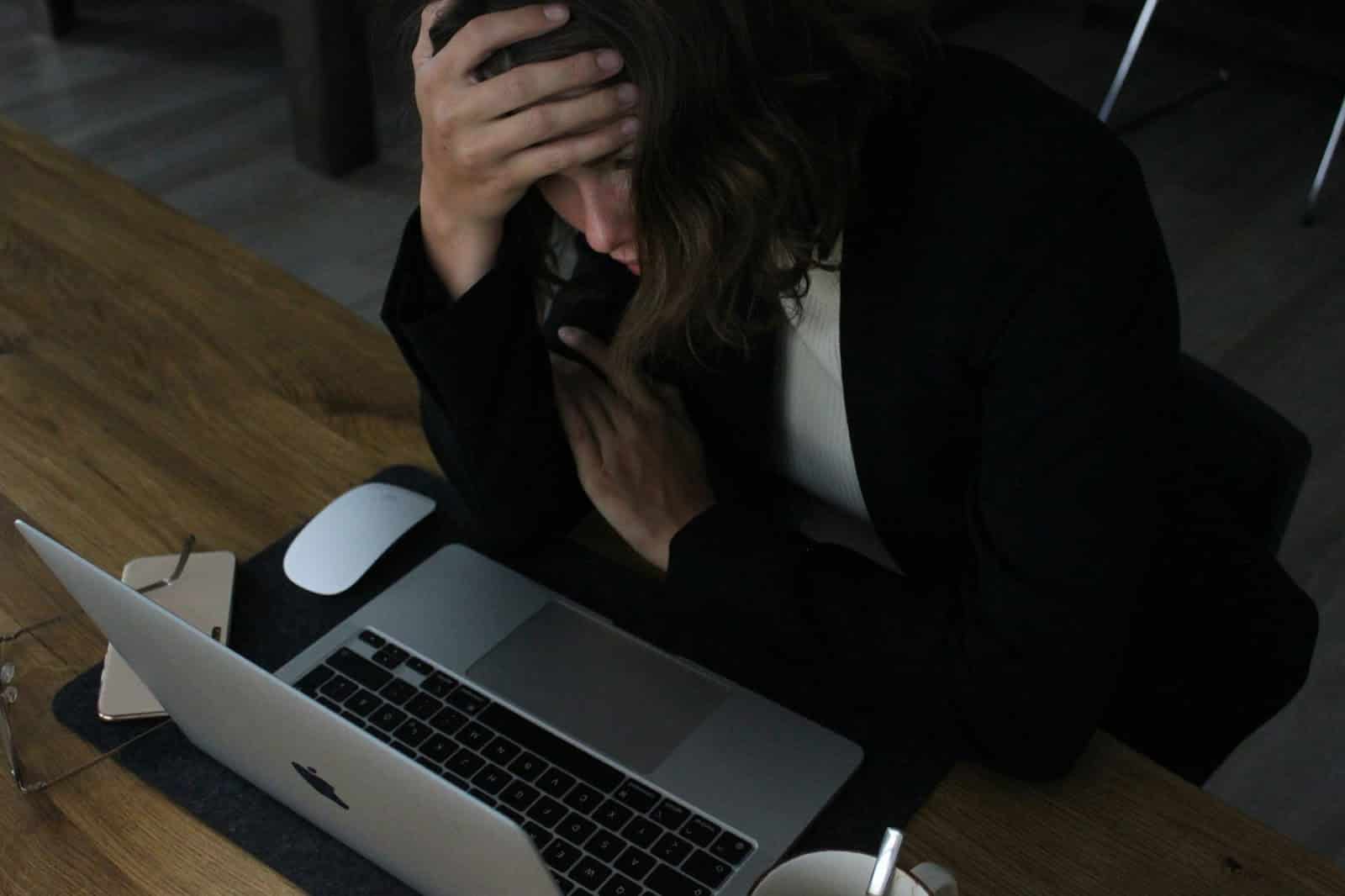 A frustrated businesswoman sitting at her desk, holding her head in her palm, looking at her laptop. She appears overwhelmed and exhausted from the impact of doomscrolling and overwork on her mental health.